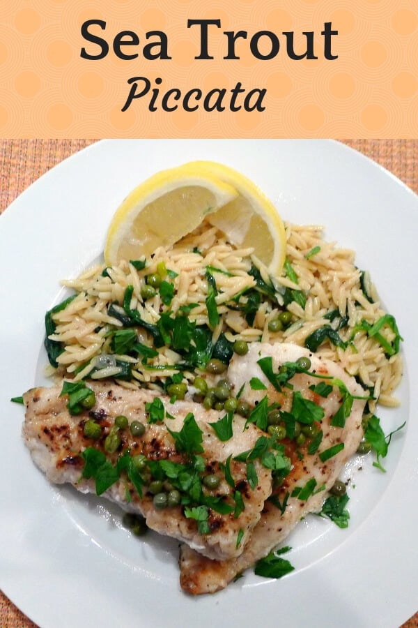 Sea Trout Piccata on a white plate with orzo spinach pasta, lemon wedges, capers, and green parsley. 