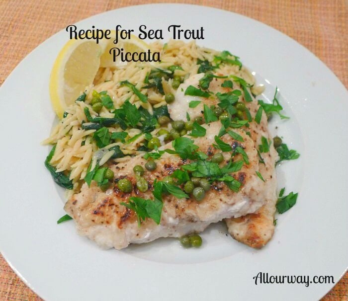 Baked sea trout piccata with lemon orzo pasta on a white plate with lemon wedges and parsley and capers over the fish. 