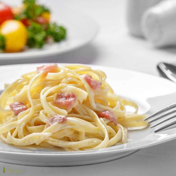 Spaghetti with pancetta bits on a white plate with a salad in background. 