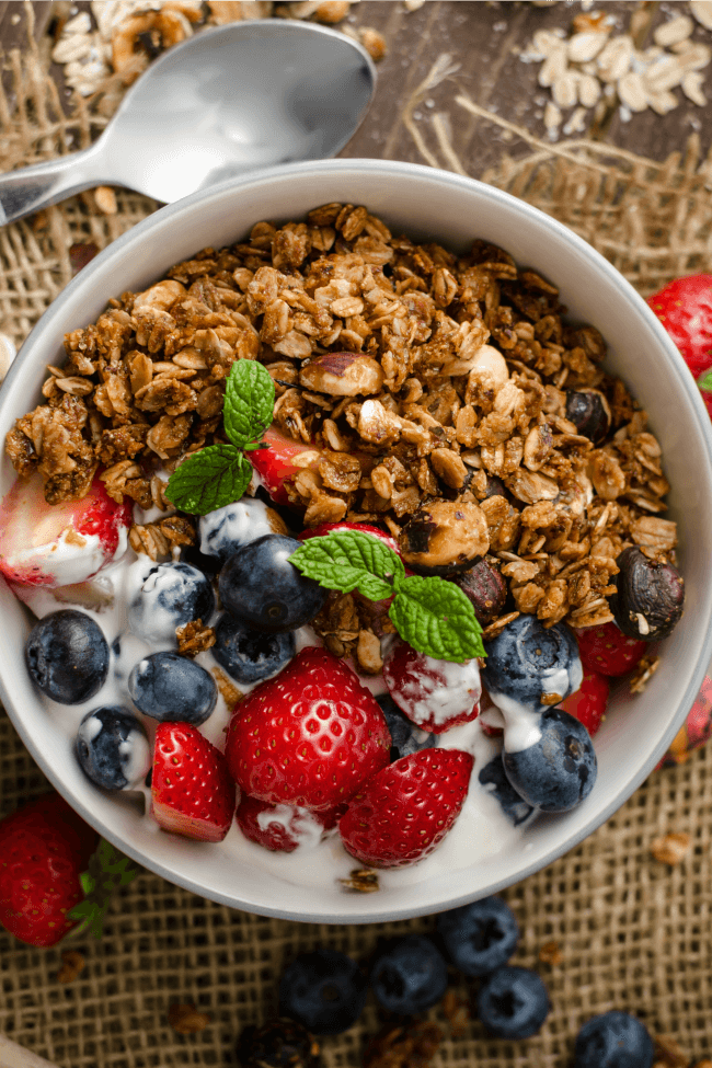 A bowl of homemade granola with blueberries and strawberries in the bowl with yogurt on the side of the granola.