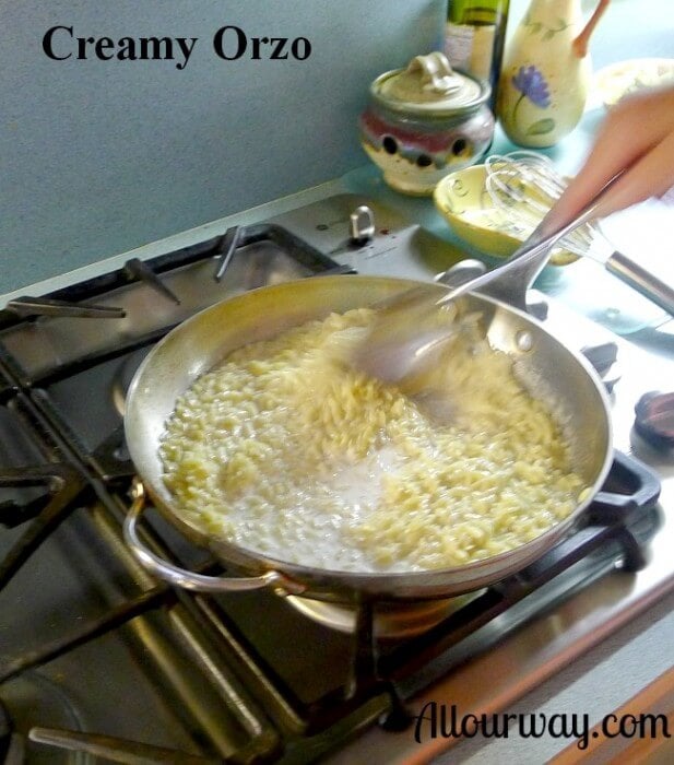 A hand with a large spoon stirring the orzo in a cream sauce in a stainless steel saucepan over a gas stove.