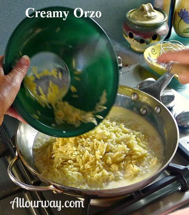 Cooked orzo in a green glass bowl poured into the sauce in the stainless steel saucepan. 