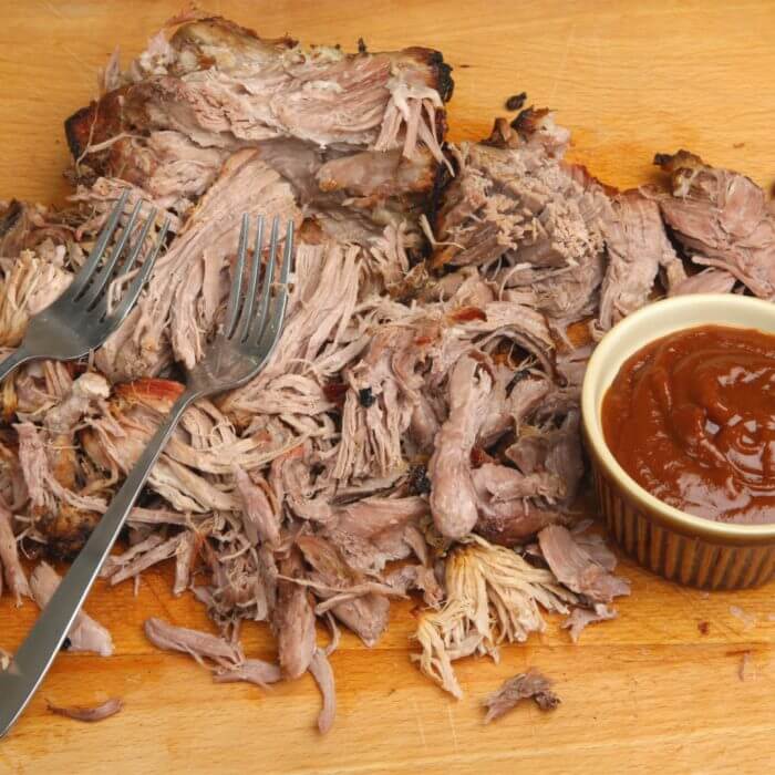 Smoked Boston Butt Recipe pork is done and shredded with forks on a cutting board with barbecue sauce on the side in a brown bowl.
