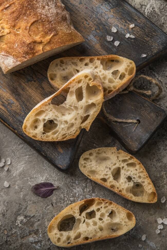 Loaf of Italian bread on a dark wood cutting board with slices of the bread on the board and counter. 