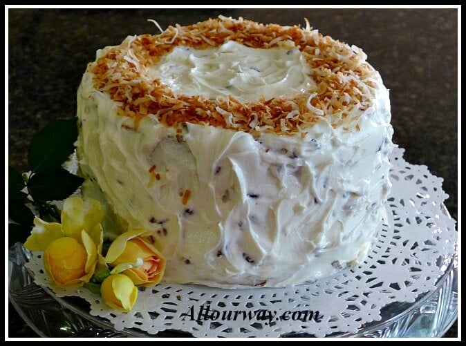 Three layer Italian Cream Cake with Toasted Coconut on a doily lined cake platter with three yellow rosebuds on the side. allourway.com