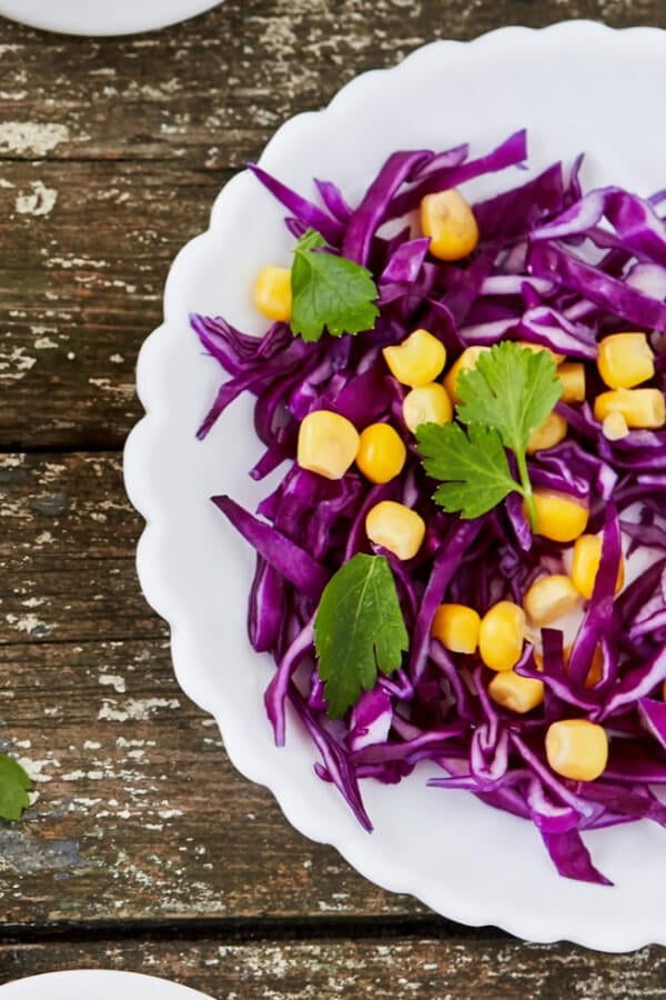 Purple coleslaw with yellow corn in a white bowl with cilantro leaves on rustic wood table. 