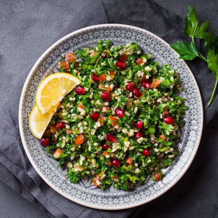 Green and red herb salad in a gray bowl with two lemon slices laying on top of a gray dishtowel that is set on a concrete surface. 