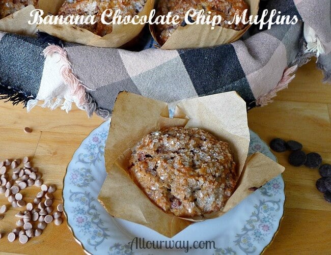 Banana Chocolate Chip Muffins with Cinnamon Chips along the side as well as chocolate chips on a wood board. Brown and black plaid cloth lined basket filled with muffins in brown parchment paper muffin cups.