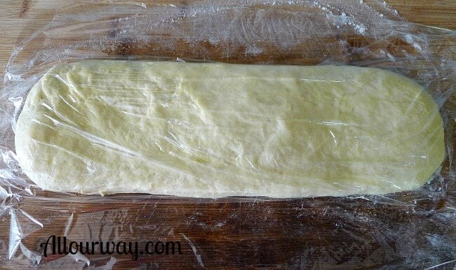 Grissini dough is shaped, brushed with olive oil and left to rest on the breadboard. Plastic wrap covers the dough. 