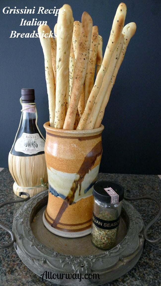 Grissini Breadsticks in an orange black and brown pottery vase. A bottle of straw-wrapped bottom chianti in the background with a shaker of Italian seasoning in the front of the tall Italian breadsticks. 