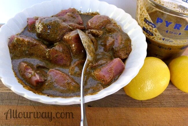 Pork cubes for kebabs marinating in brown marinade in a white bowl. Lemons are on the side and a spoon is in the bowl for turning the meat. 
