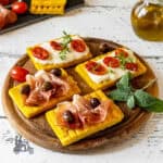 A wooden board holds four grilled polenta crostini with cheese and Italian meats.
