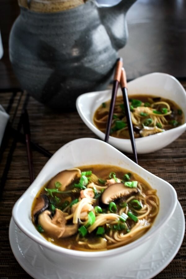 Two white triangular bowls filled with hot and sour soup. Gray teapot in background with two chopsticks. 