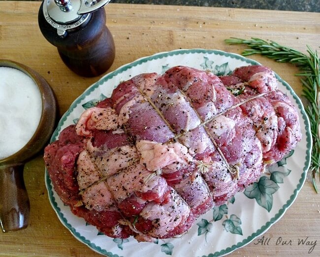 Leg of lamb tied and seasoned with pepper on white and green platter with salt and pepper mill on the side. Sprig of fresh rosemary is next to platter. 