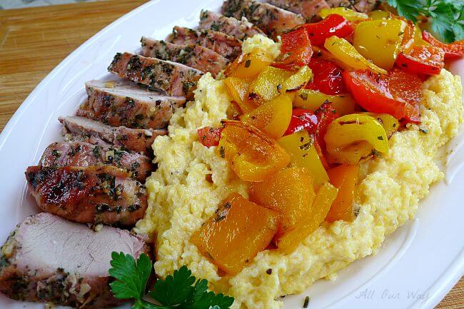 Grilled whole pork tenderloin slices with colored peppers over polenta on a white platter @ allourway.com