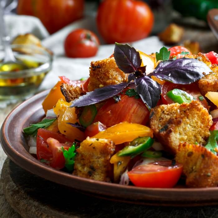 a close up of panzanella an Italian tomato bread salad in brown bowl with purple basil on top and tomatoes in background. 
