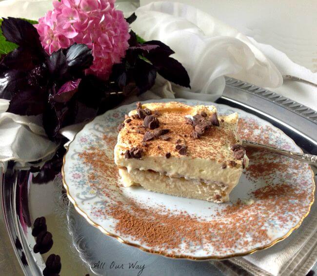 Tiramisu Cake square on a china plate with gray, red, and blue flowers and a silver fork on the side. A pink hydrangea is behind the cake serving. 