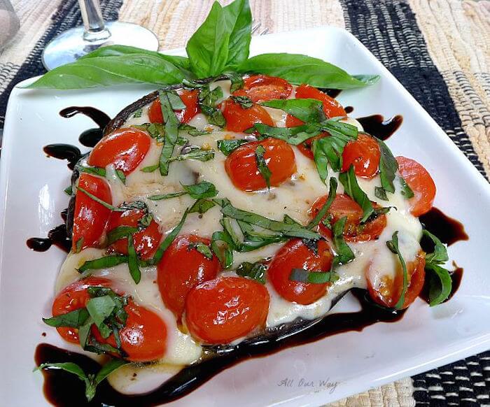 A grilled large portobello mushroom stuffed with mozzarella cheese, halved grape tomatoes, and slivers of basil set on a square white plate with a drizzle of dark balsamic vinegar all around the plate.