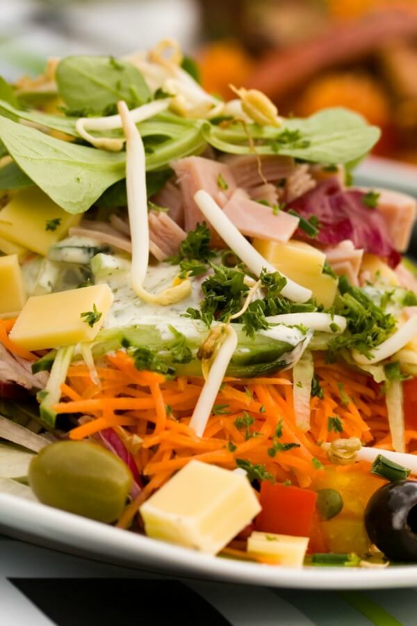 A chef salad closeup with salad greens, carrots, olives, cheese, and sliced meats, tomatoes. 