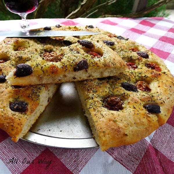 Focaccia Pugliese with cherry tomatoes and kalamata olives sliced into a wedge over a red and white checked tableclth an a glass of red wine on the side. @allourway.com