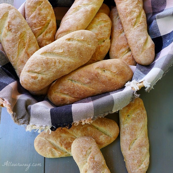  Sandwich Rolls fill a brown and black plaid napkin lined basket with several rolls on a gray wood background. 