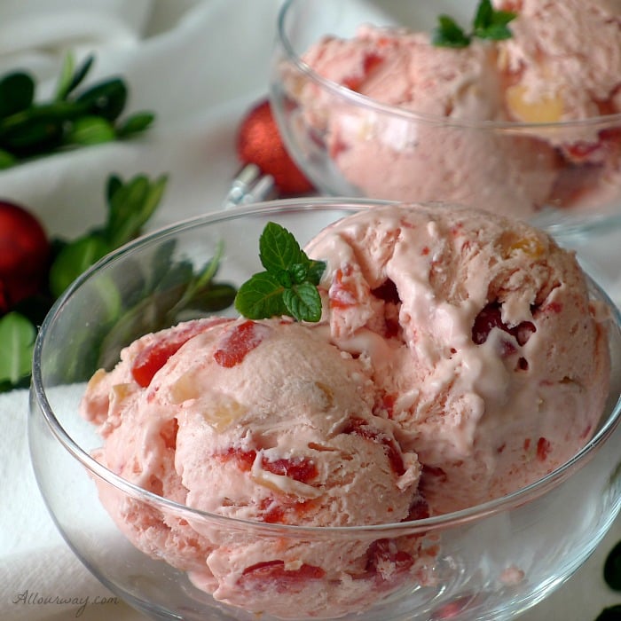 Two scoops of pink Cherry Almond Amaretto No-Churn Ice Cream in a delicate glass bowl surrounded by red mini Christmas balls and green boxwood on a white snowy background. 