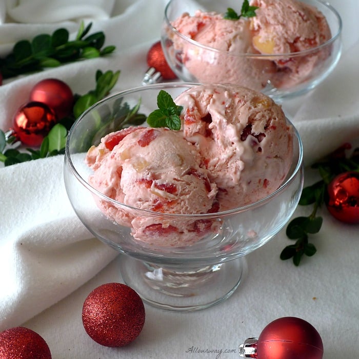Two Pink scoops of Cherry Almond Amaretto No-churn ice cream with a sprig of mint in two glass parfait bowl with mini red christmas balls and green boxwood surrounding the bowls. 