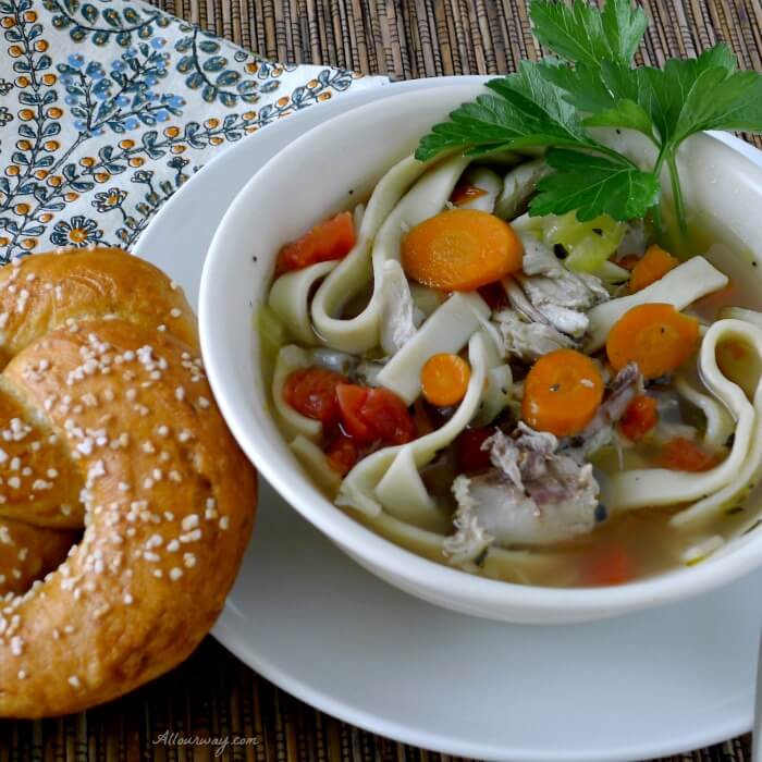 Homemade Chicken Noodle Soup served in a white oversized cup with a Homemade Pretzel next to it and a black and blue print napkin next to it. @allourway.com