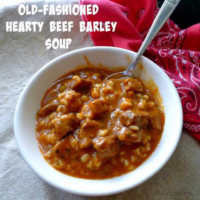 Close up of deep red Beef Barley Soup in a white bowl with a spoon stuck in the center. A red bandana is on the right side. 