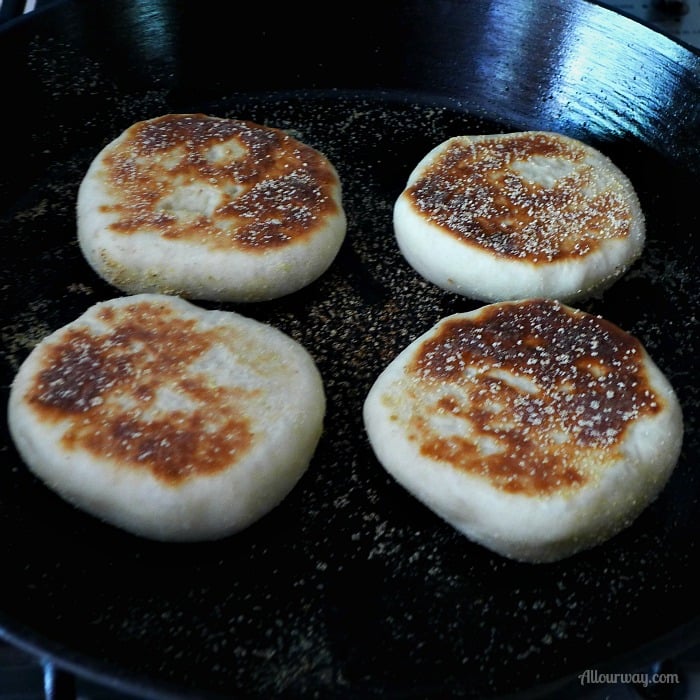 English Muffins cooking in a black cast iron skillet. They are already browned on one side. 