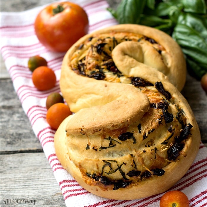 On a white and red striped dish towel is Herb Cheese Swirl Pane Italiano - Bread stuffed with a savory filling. Tomatoes are on the side with fresh basil in the background. @allourway.comm