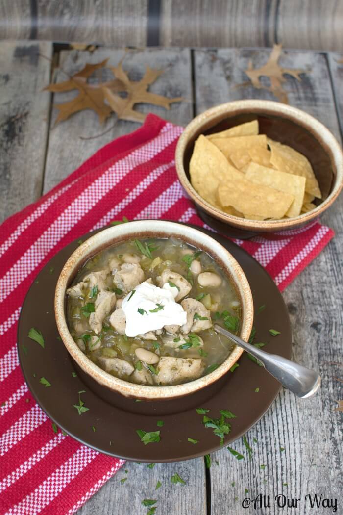 Wickedly Good Spicy White chicken Chili with lime and cilantro in brown glazed bowl with a dollop of sour cream on top. Tortilla chips in another brown bowl and they are set atop a red and white striped tea towel on rustic gray boards with brown oak leaves strewn around. 