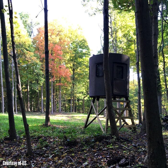 A cylindrical dark brown deer-blind on stilts in the middle of a clearing in the woods with leaves turning colors in autumn. 