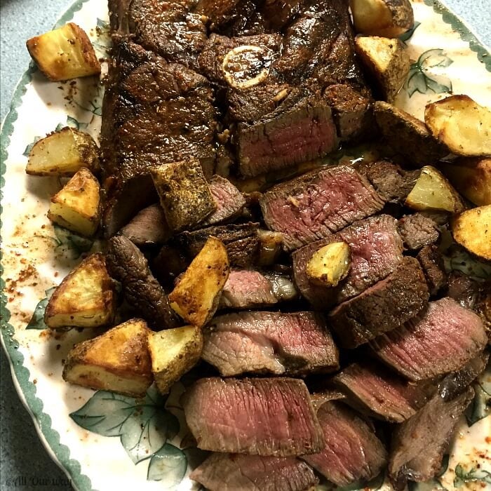 Close up of a grilled Venison Steak surrounded by brown potato wedged on a green and white plate with leaf pattern.