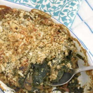 Spoon in a gratin dish filled with spinach gratin and blue floral tea towel on side.