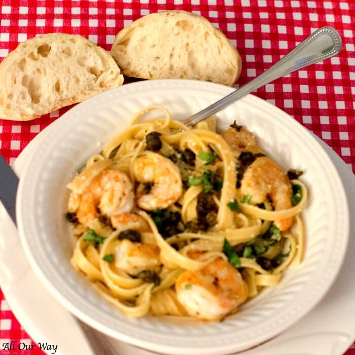 Seafood pasta dish flavored with parmesan, basil, and capers in a white bowl with a red gingham tablecloth. 