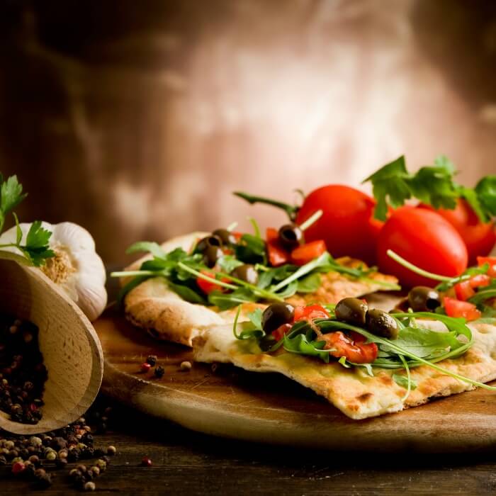 Flatbread made into a pizza with arugula, tomato and olives on top. Bulb of garlic on the side along with a scoop of peppercorns.