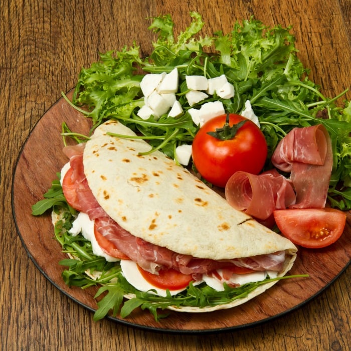 Flatbread folded in half is filled with prosciutto, fresh mozzarella cheese, tomatoes and arugula. All on a wooden server with extra tomato, prosciutto, mozzarella and arugula on the side. 