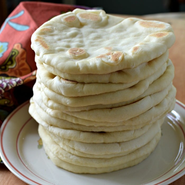 Flatbread is stacked up one on top of each other on a white plate with russet edge. A brick colored napkin with turquoise and gold is on the side.