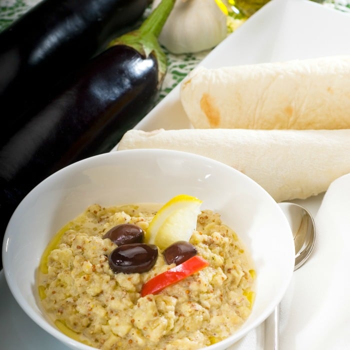 A white bowlful of baba ghanoush with 3 black olives on top, a slice of red pepper and a sliver of lemon. To the upper left are two eggplants and on the side of the white bowl are two flatbreads rolled up.