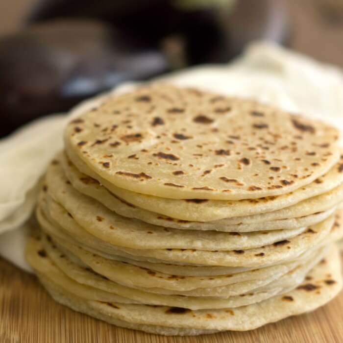 A stack of cooked round flatbread on a wooden cutting board with a white cotton towel in the background.