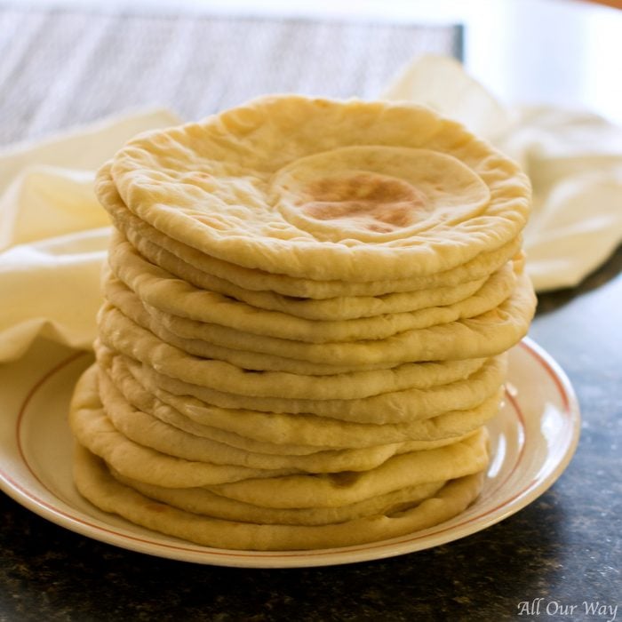 On top of a russet rimmed plate is a tall stack of pancake like flatbread. A white cotton towel is in the background.