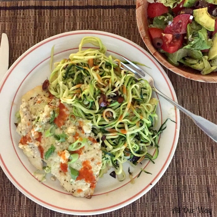 Parmesan Broiled Flounder on a white and brick colored plate with zucchini almond noodles. Plate is on a brown placemat with a wooden bowl filled with a tossed salad.