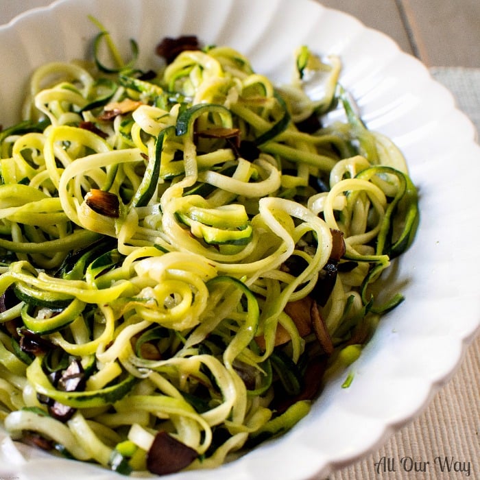 Long strands of zucchini noodles and almonds in a white fluted bowl on top of a green and white placemat.