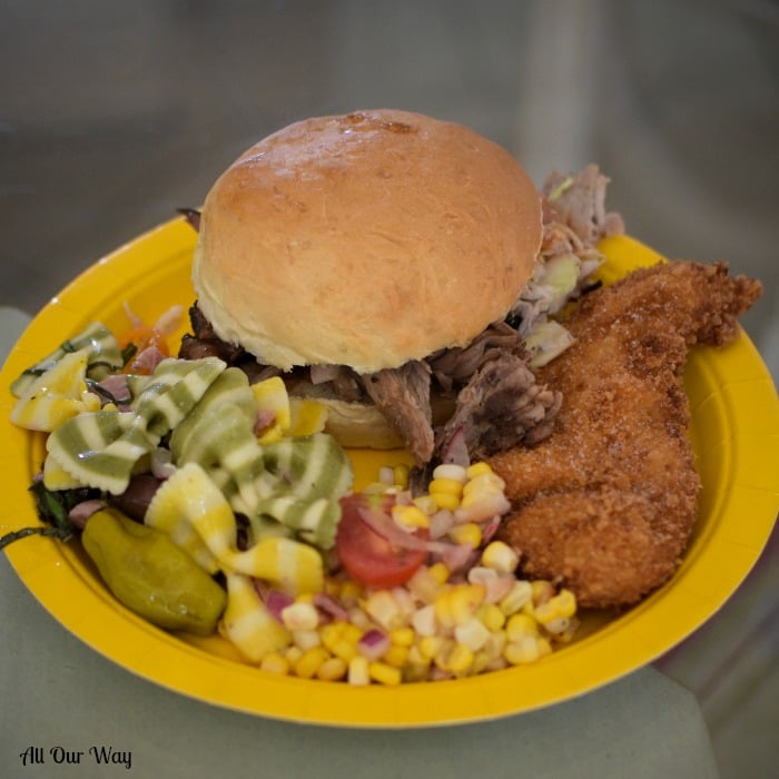 A yellow picnic plate holding pasta salad, corn, fried chicken, and a pulled pork sandwich. 