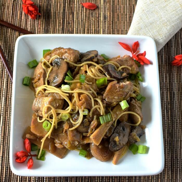 Chop Suey is a Chinese-American Stew with thin pasta and sprinkled with green onions on a white square plate with red salvia blossoms sprinkled around the plate and brown bamboo placemat with a tan linen placement on the side. 