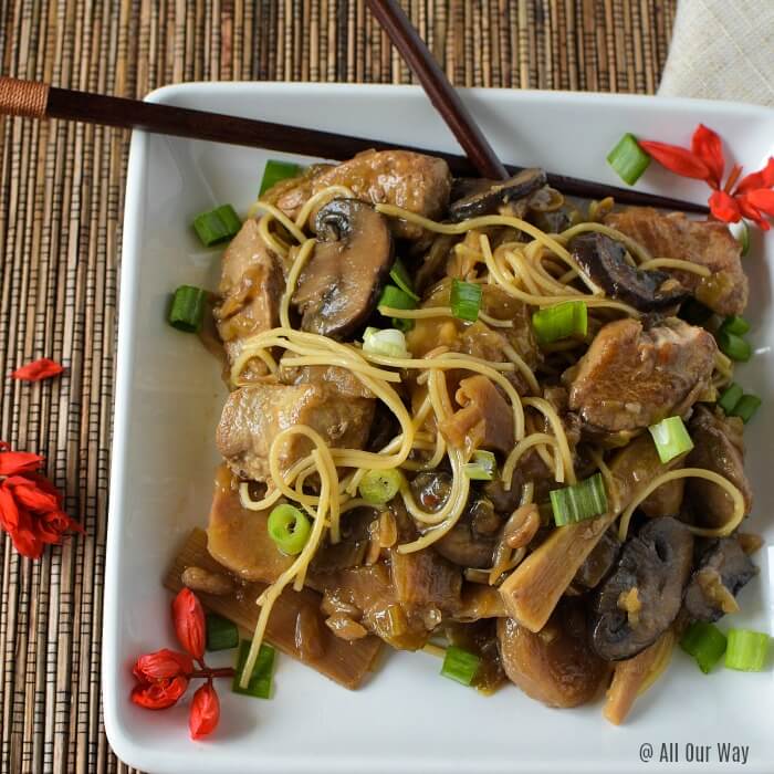 Close up of Chop Suey a Chinese-American stew in a white square plate with bamboo chop sticks and green onions garnishing the dish with red salvia blossoms on placemat and dish.