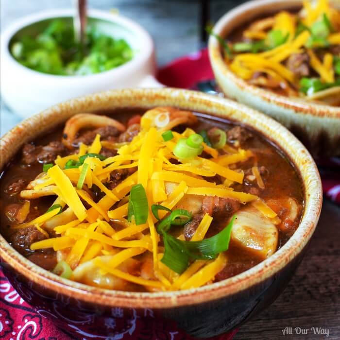 Two brown glazed bowls filled with hearty venison chili that's topped with shredded yellow cheese and green onion rings. Small white bowl of green onions in background. 