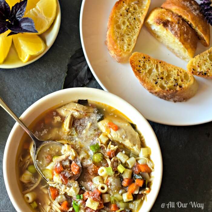Bowl of chicken vegetable soup with spoon, lemon wedges, and bread slices 
