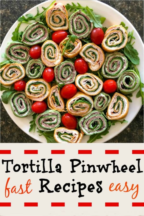 A large white platter filled with colorful spinach and red pepper tortillas and green arugula and grape tomatoes in between the round pinwheels. 